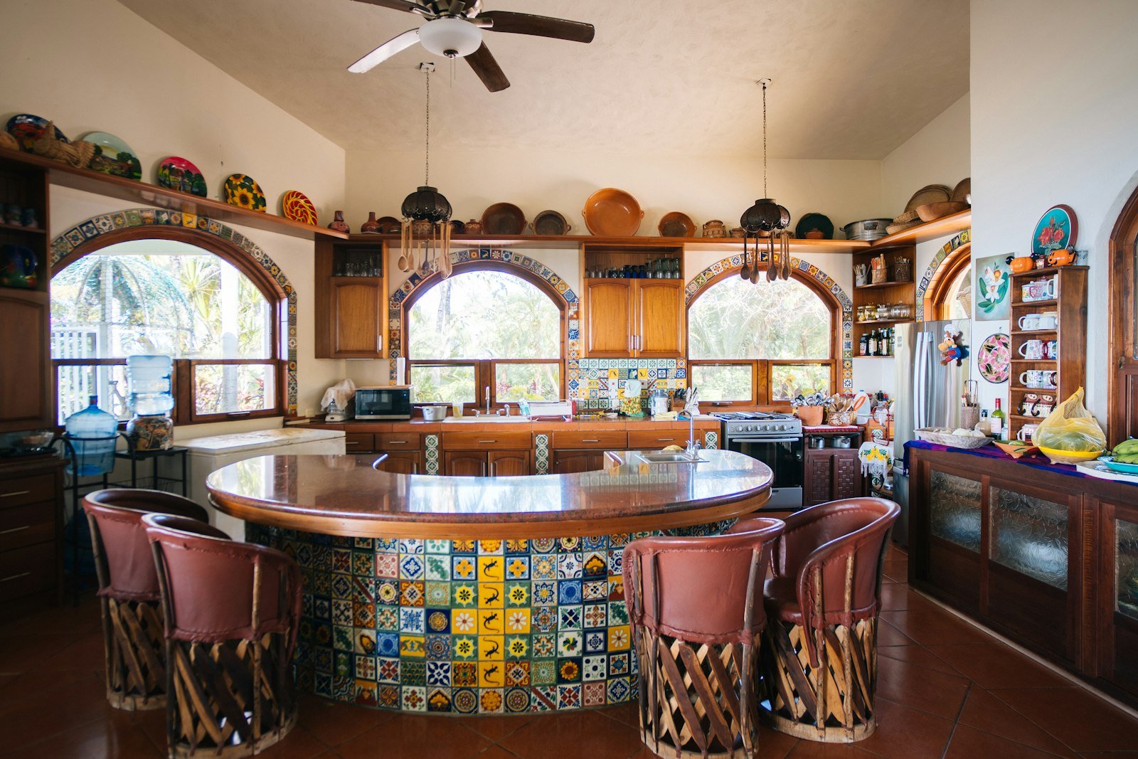 Kitchen with a round island covered in multicoloured patterned tiles, pink bar stools and shelves of colourful pottery under arched windows