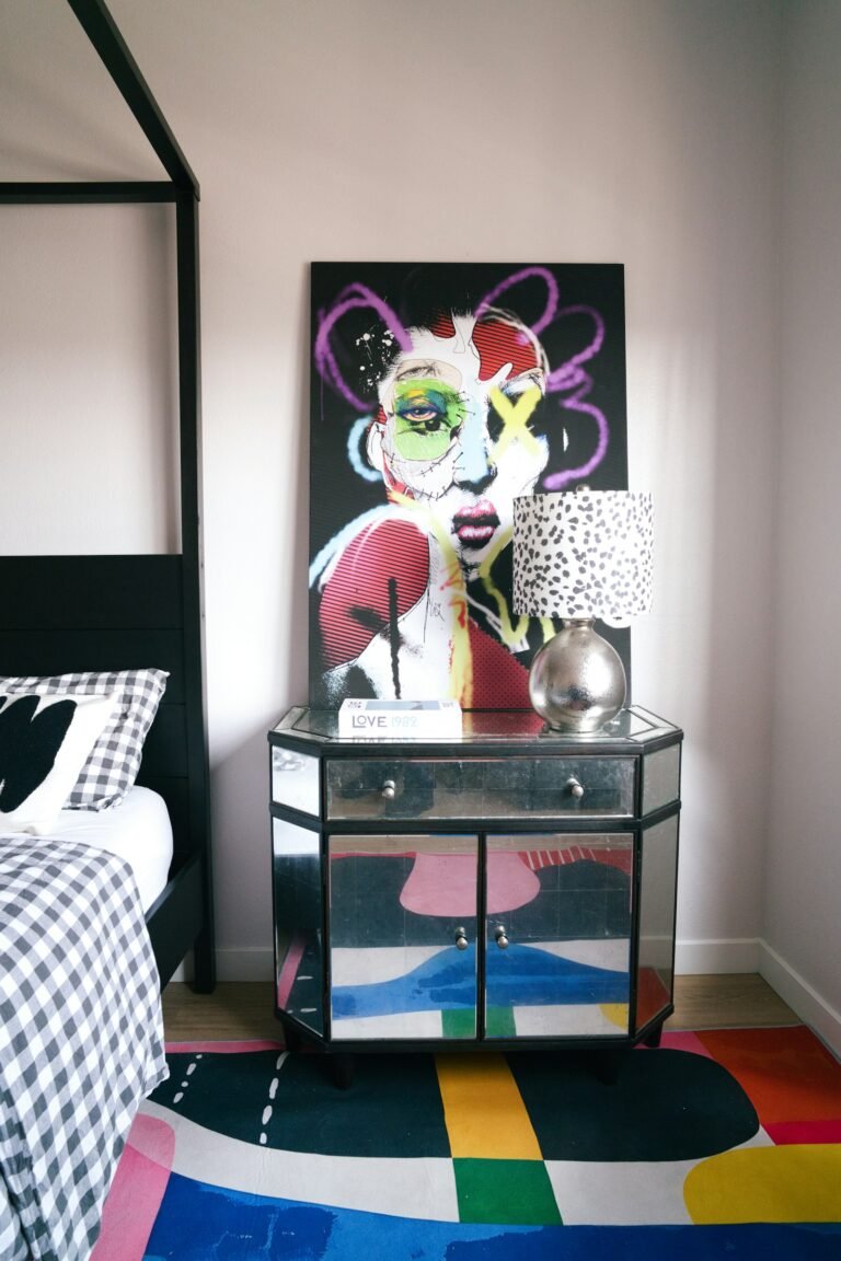 Bedroom corner with a large colourful pop-art portrait above a mirrored nightstand, a patterned lamp and a checked duvet