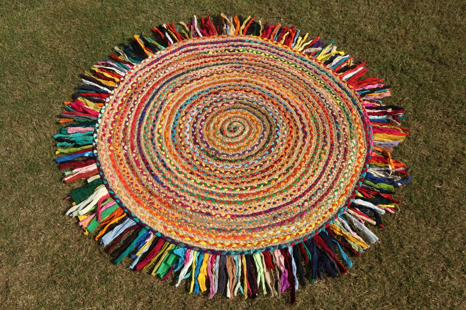 Round braided rug made of multicoloured fabric strips with a long rainbow fringe, lying on grass