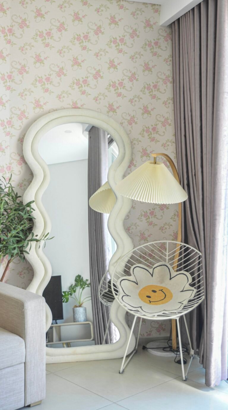 Tall mirror with a wavy white frame beside a pleated lamp and a woven chair in a softly lit room